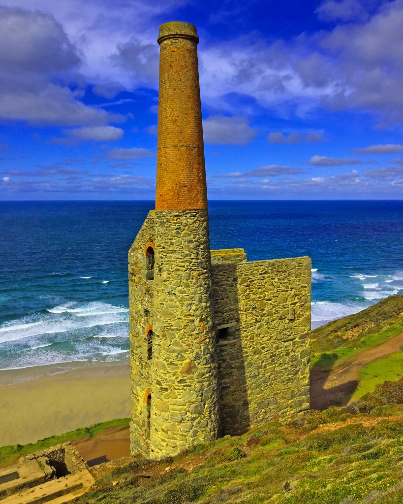mine chimney and building in cornwall by beach and sea with blue cloudy sky