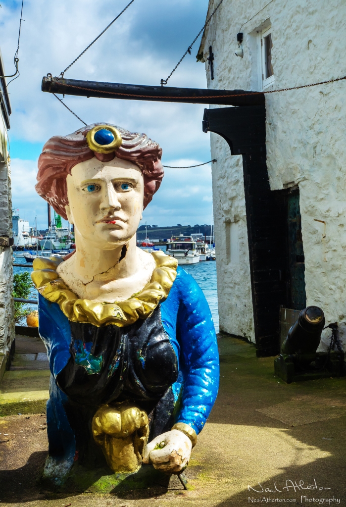 A ships figurehead rests in a small alleyway in front of a harbour with old stone buildings either side