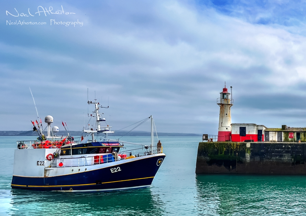 A fishing boat swings by a lighouse at the entrance to a fishing port under a cloudy sky