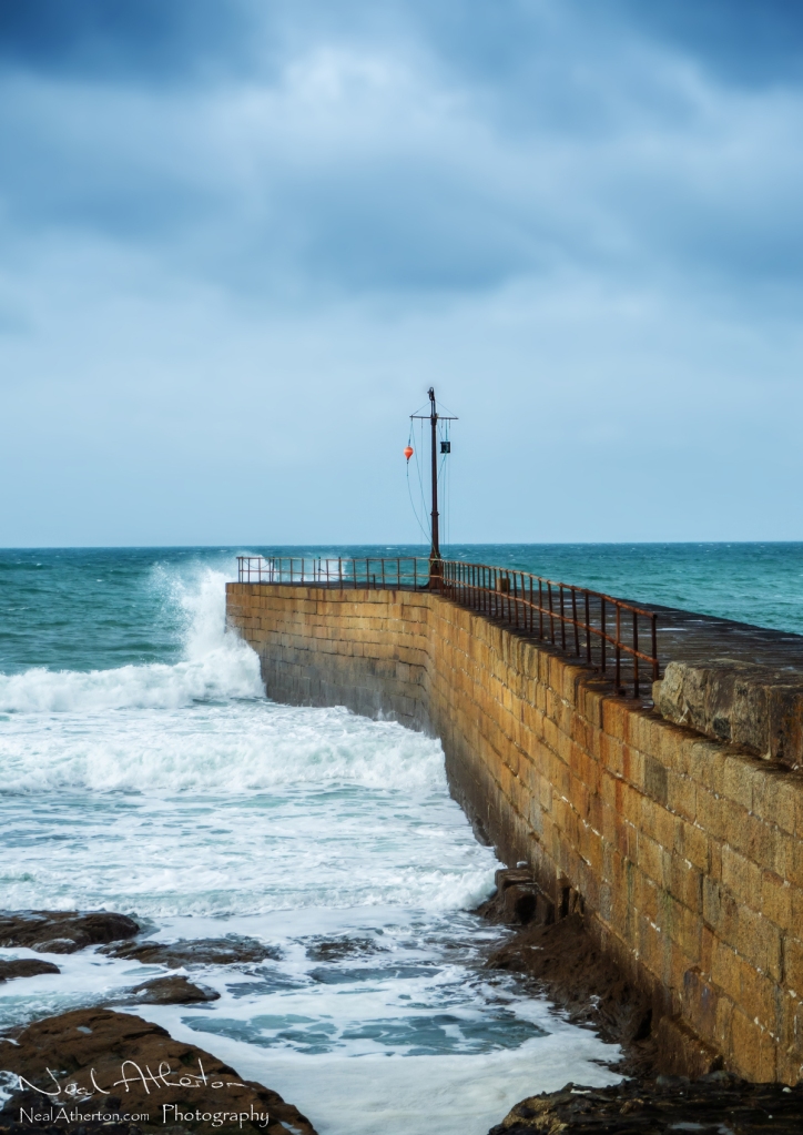 Waves hit a long stone pier with a warning bouy raised on a metal pole