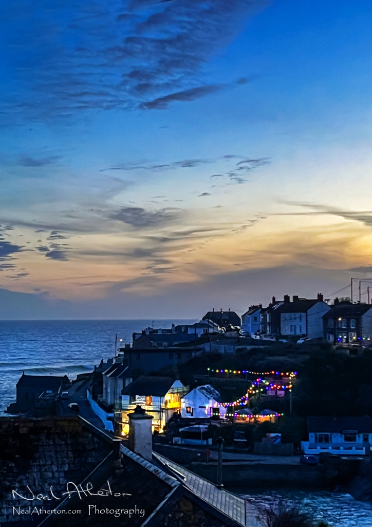 In evening dusk light a pub is lit up by rows of colured lights under the clouds of a sky after sunset with a harbour in front of the pub
