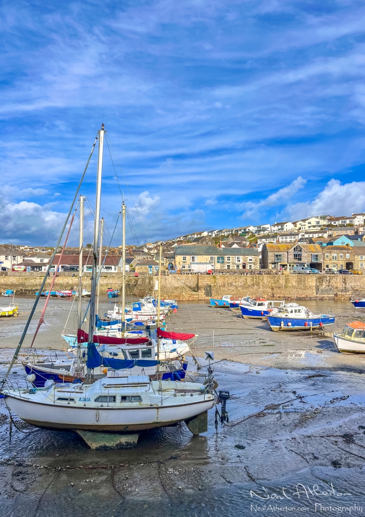 row of yachts in a harbour in Cornwall at low tide with a pub opposite and house rising up a hillside under blue sky