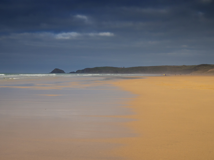 A wide sandy beach with a treatening dark sky with cliffs ans waves coming in